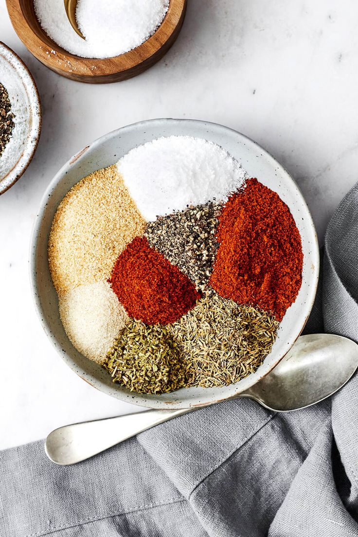 spices and seasonings in bowls next to spoons on a white counter top with gray linen