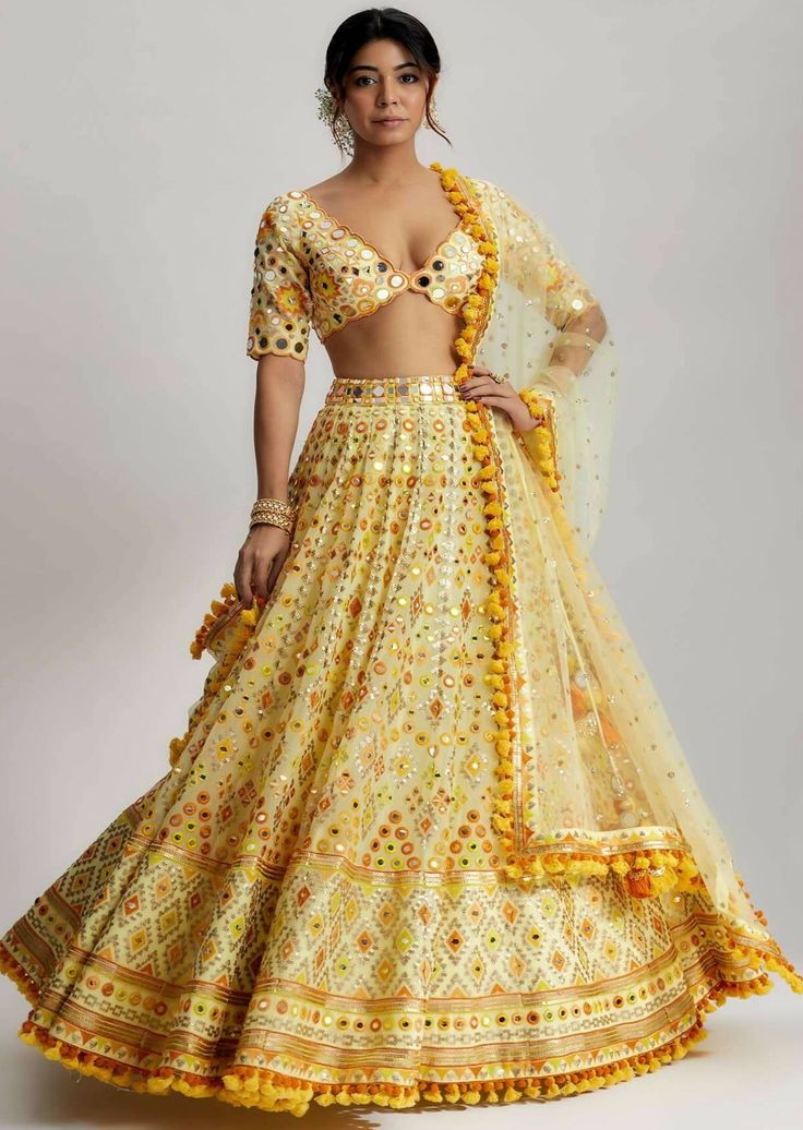 Adorned with playful pompoms and tassels, the anha lehenga exudes a playful charm. These charming embellishments add a whimsical element to the ensemble, enhancing its overall appeal. The carefully selected fabric drapes gracefully, accentuating the beauty of the wearer and creating a regal silhouette. Bohemian Embellished Wedding Sets, Bohemian Embellished Lehenga For Weddings, Bohemian Embellished Wedding Lehenga, Traditional Dress With Tassels For Diwali, Bohemian Lehenga With Mirror Work For Reception, Bollywood Choli With Tassels, Festive Choli With Tassels, Bohemian Embellished Lehenga With Traditional Drape, Yellow Bohemian Lehenga For Festive Occasions