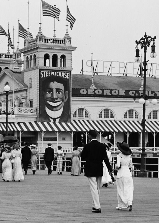 black and white photograph of people walking on boardwalk in front of building with american flags