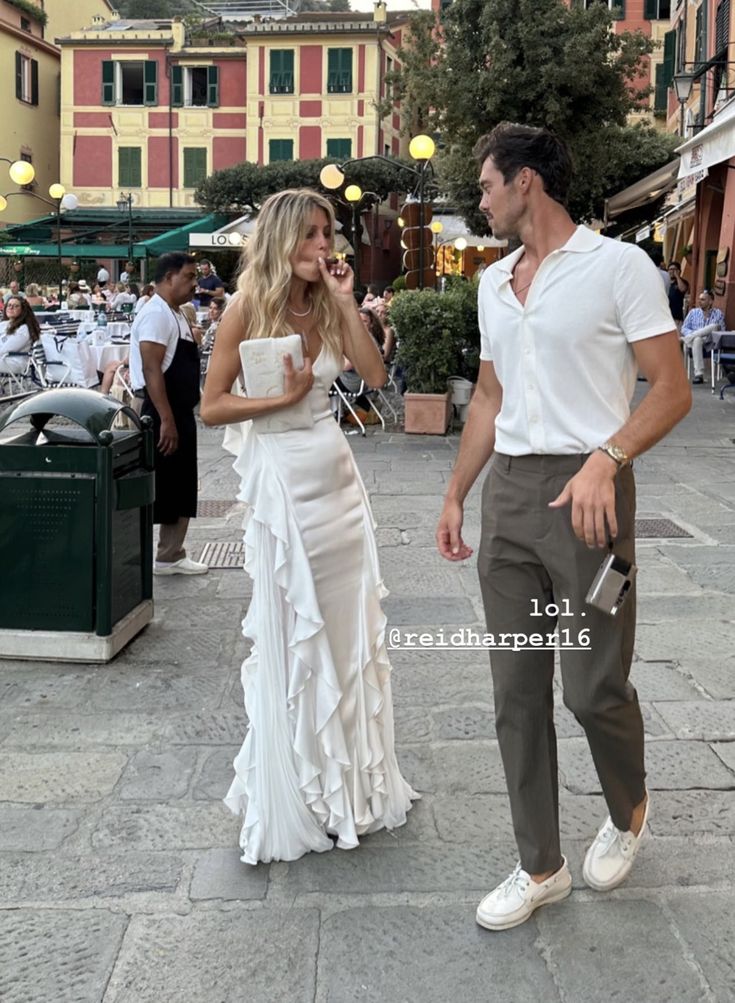 a man standing next to a woman in a white dress on a city street with buildings behind her