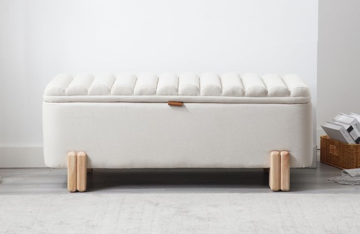 a white bench with wooden legs in front of a white wall and basket on the floor