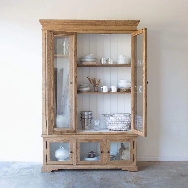 a wooden cabinet with glass doors and dishes on the top, in front of a white wall