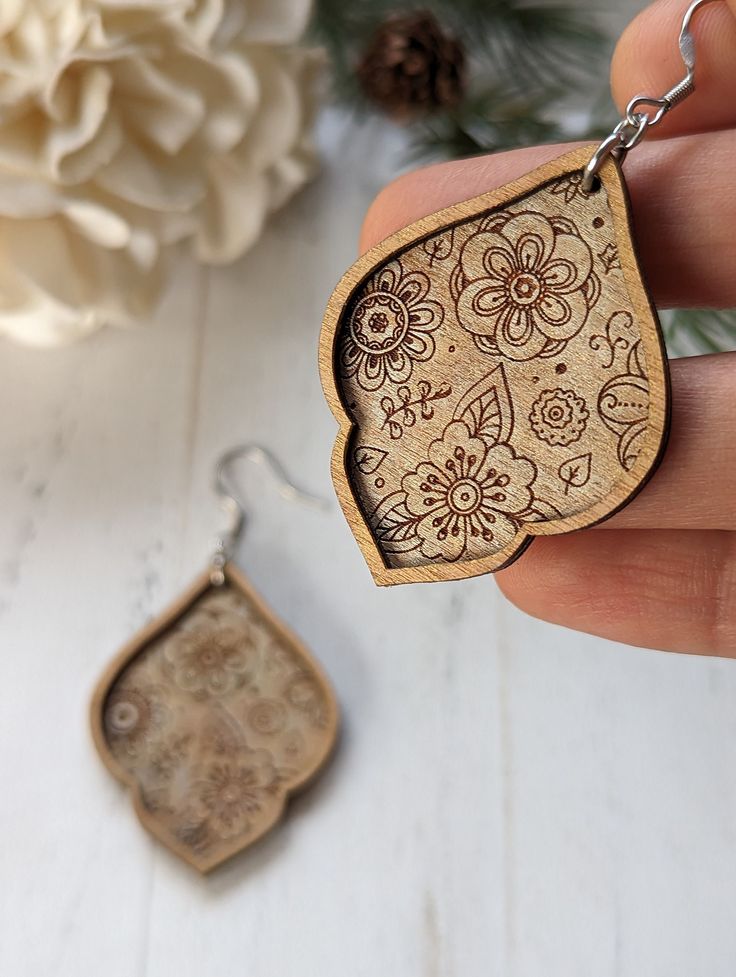 a hand holding a wooden keychain with an ornate design on it and a flower in the background