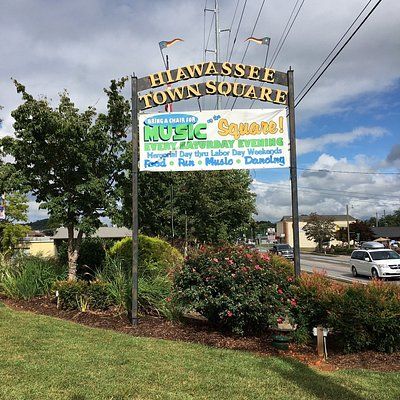 a sign that is in the grass next to some bushes and trees with cars parked on it