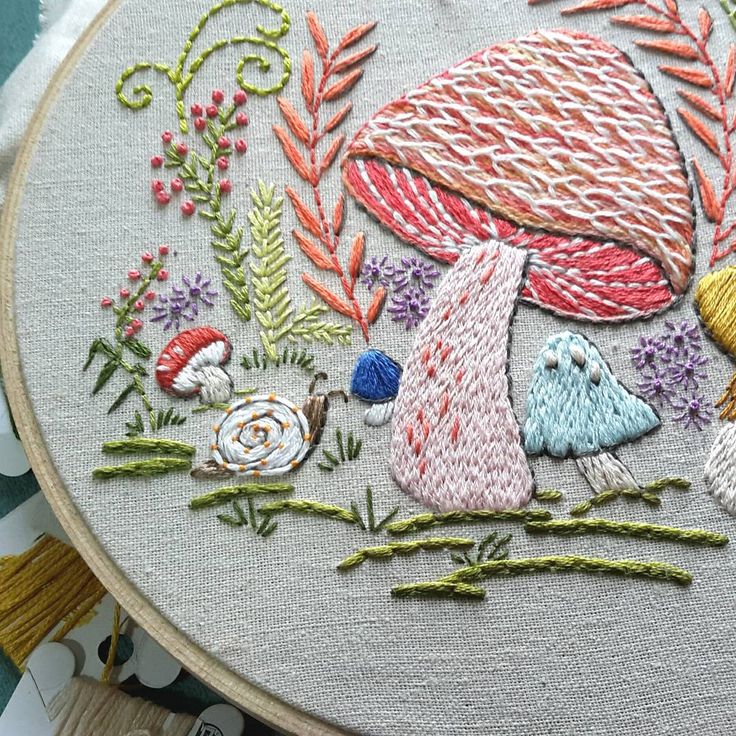 a close up of a embroidery on a hoop with flowers and mushrooms in the background