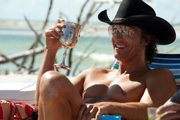 a man wearing a cowboy hat and glasses holding a wine glass while sitting on the beach