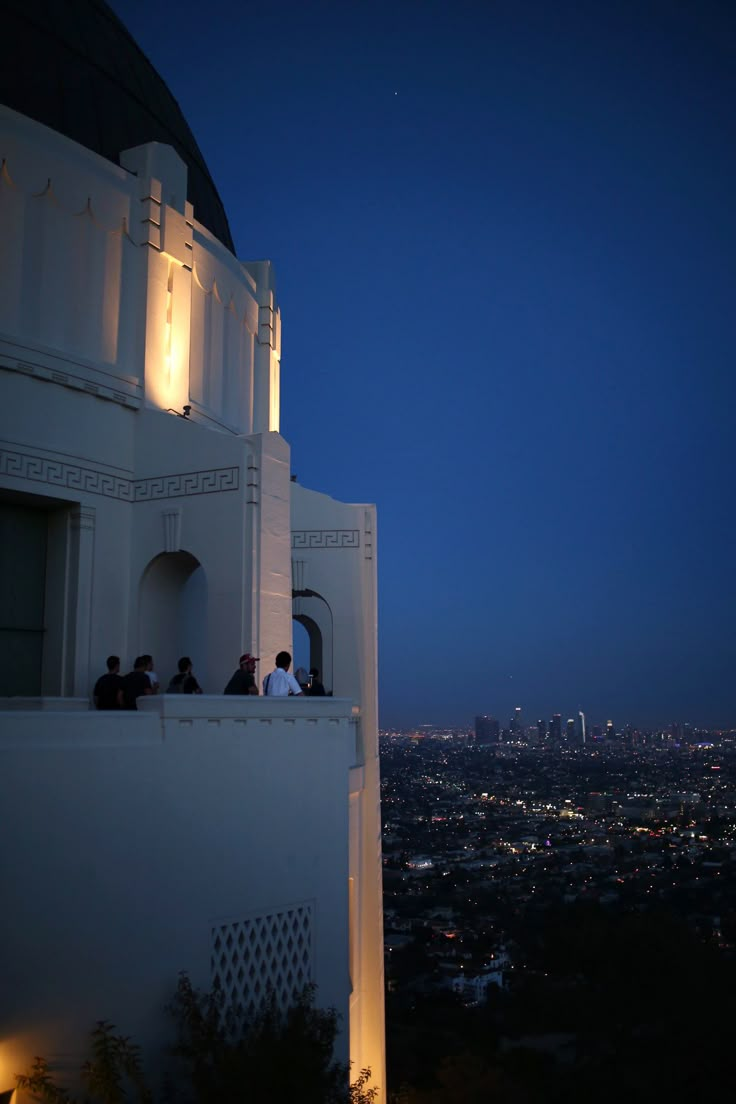 people are sitting on the top of a building at night with city lights in the background