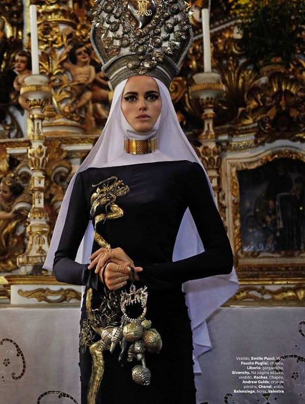 a woman dressed in black and gold with a crown on her head, standing next to an ornate wall