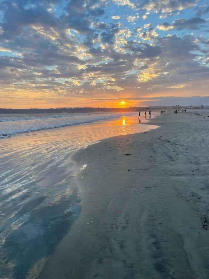 people are walking on the beach as the sun goes down in the distance with clouds