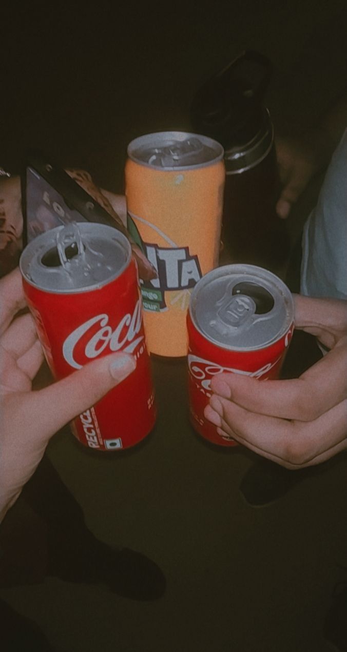 three people holding cans of coca - cola on top of a table with other drinks