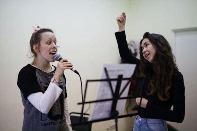 two young women singing into microphones in front of music sheets and sheet music stands