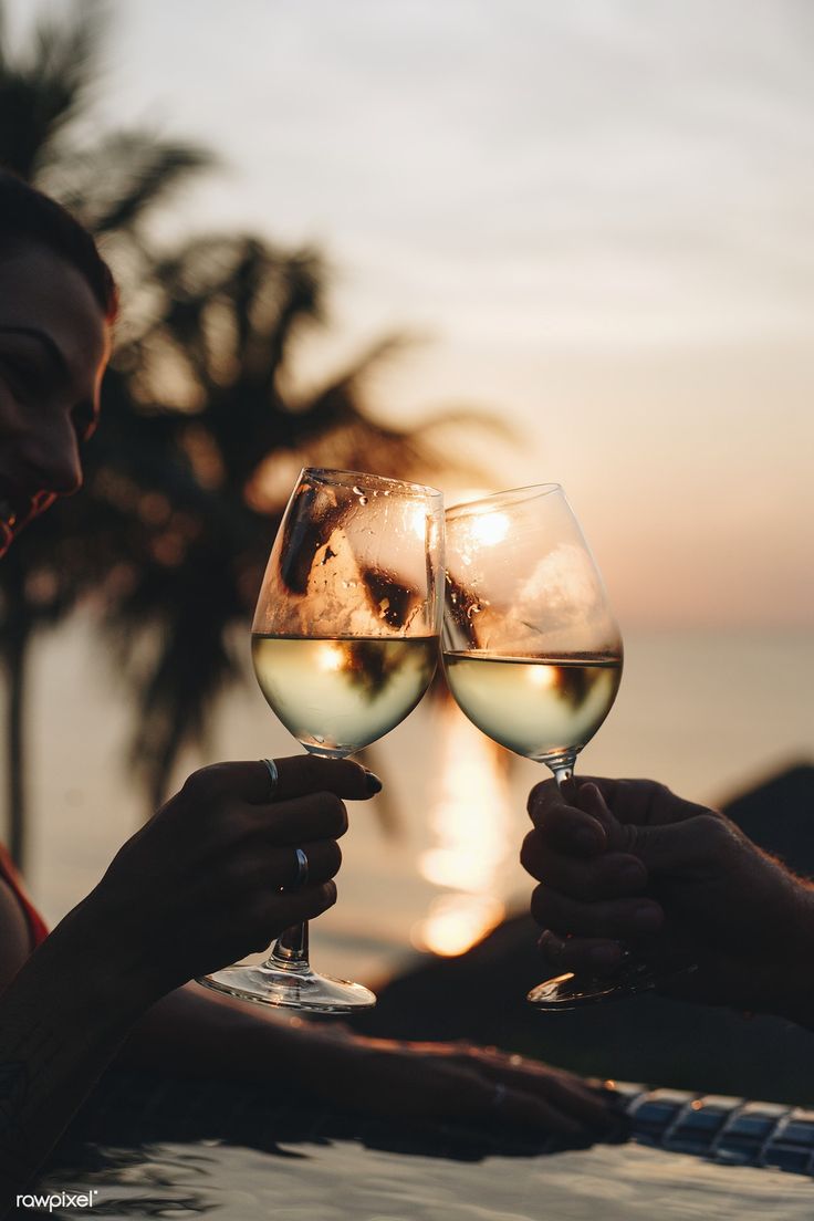 two people toasting with white wine in front of the ocean and palm trees at sunset