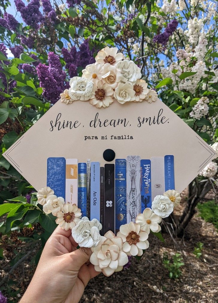 someone holding up a sign that says shine dream smile with flowers around it and books in front