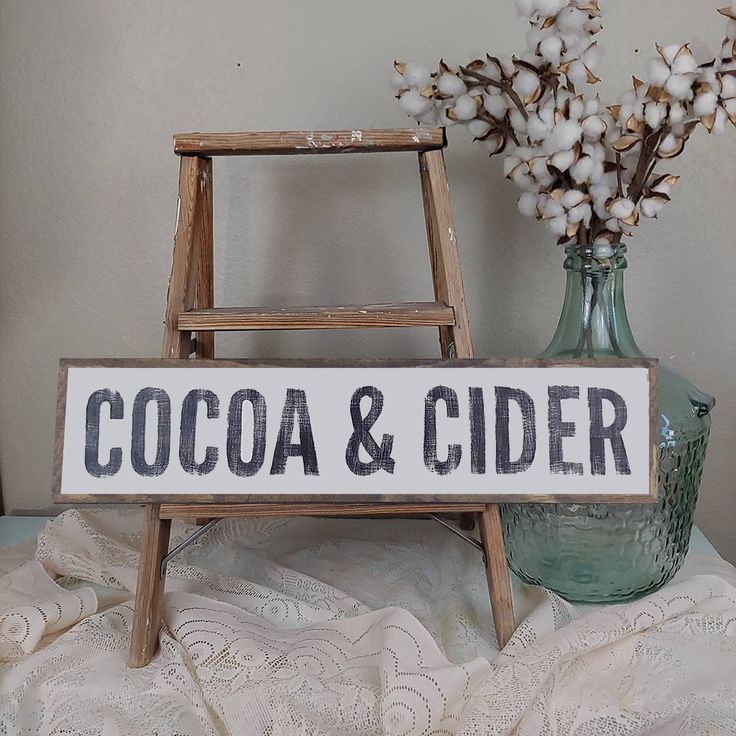 a wooden sign that says cocoa and cider next to a vase with cotton flowers