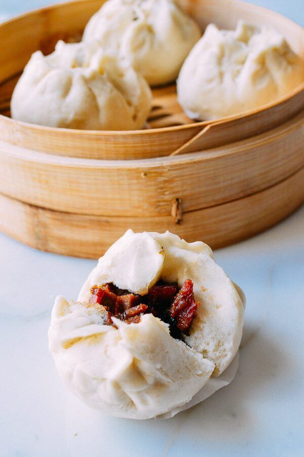 three dumplings in a bamboo basket on a white countertop next to a wooden bowl