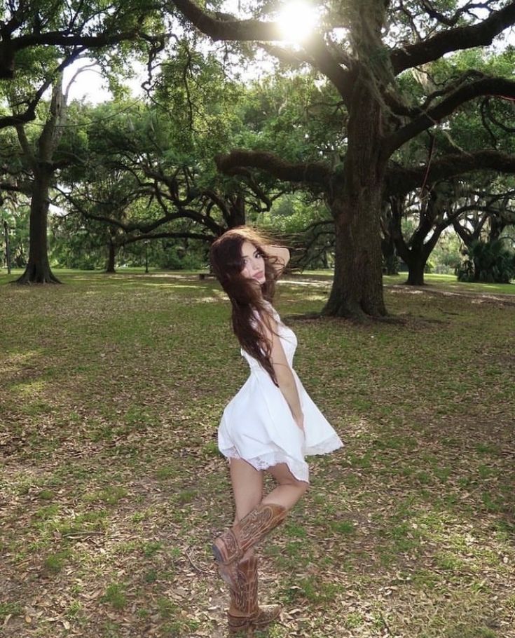 a woman in a white dress and cowboy hat posing for the camera with trees in the background