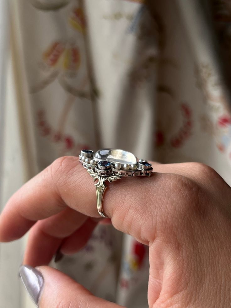 “Art Deco” was an era marked by extravagance and innovation. The Art Deco style itself was an eclectic mix of heavy geometric shapes, with curved ornamental elements. The resulting silhouettes were often monolithic but elegant, with applied decorative elements. This Moonstone and Sapphire Edwardian Conversion Ring is a wonderful example of this iconic Art Deco style. Crafted in 20 carat white gold, this ring immediately captures the gaze with its dazzling array of miniature pearls and sapphire stones haloing a truly luminescent cabochon moonstone. What makes this treasure particularly special is that it is a conversion piece; the beautiful moonstone center was once part of an antique brooch. Now, with the use of a 14 carat gold replica shank, it has been transformed into the glorious ring Luxury Open Ring With Unique Design, Luxury Polished Moonstone Ring, Luxury Sapphire Ring With Intricate Design, Luxury Oval Jewelry With Ring Detail, Luxury Oval Ring With Detailing, Elegant Polished Topaz Open Ring, Luxury Jewelry With Unique Design For Wedding, Art Deco Multi-stone Oval Sapphire Ring, Art Deco Oval Multi-stone Sapphire Ring