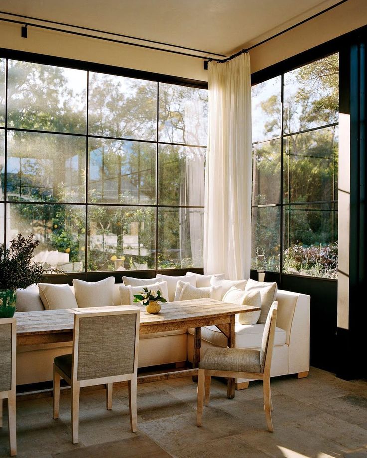 a living room filled with furniture and large windows covered in white draping next to a wooden table