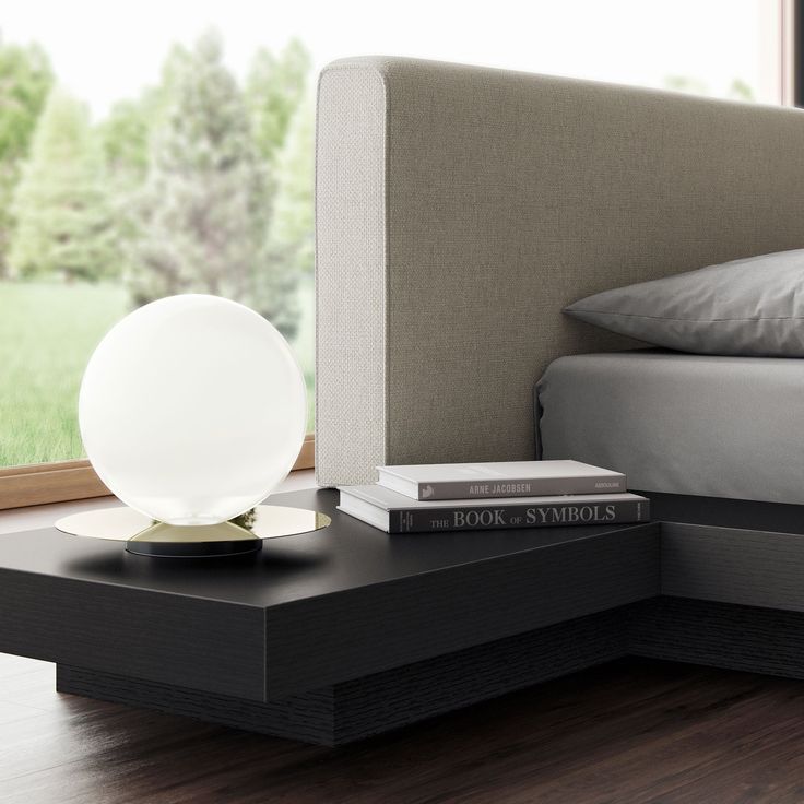 a nightstand with books and a lamp on it in front of a window overlooking a bed