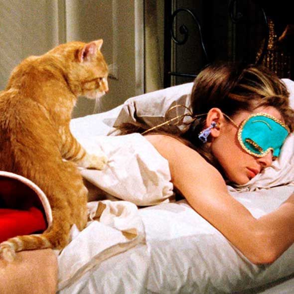 a woman laying in bed with a cat on her head and sleeping mask over her eyes