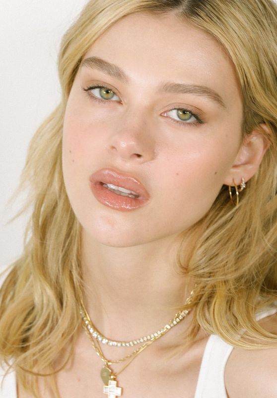 a woman with blonde hair wearing a white tank top and gold necklace, looking at the camera