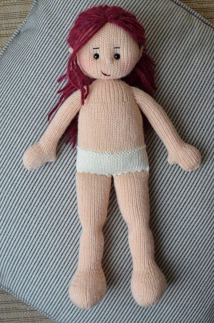 a pink knitted doll laying on top of a gray and white striped pillow with red hair