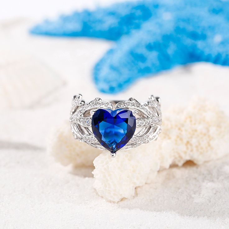 The coral evokes memories of your favorite days at the beach as well as evenings beneath the celestial sky. Created in sterling silver, it features a blue heart cut stone at the center, while two corals wrap the center stone in a sparkling embrace and continue into the ring's shank.  Our coral inspired ring allows you to carry a tangible reminder of your favorite travels and adventures indefinitely.Carat Weight: 5.2 ctStone Size: 9*9 mmStone Type: Jeulia® StoneNumber of Stones: 1 Stone Color: Sapphire BlueStone Shape: HeartCarat Weight: 1.435 ctStone Size: 1.5,1.3,1.1 mmStone Type: Jeulia® StoneNumber of Stones: 110 Stone Color: Diamond WhiteStone Shape: RoundWeight: 6 gWidth: 12.5 mmHeight: 7.3 mmThickness: 3 mmMaterial: 925 SilverPlating Color: Silver Blue Ocean-inspired Jewelry For Anniversary, Ocean-inspired Blue Jewelry For Anniversary, Fine Jewelry Blue Heart Ring For Valentine's Day, Blue Heart Ring For Valentine's Day, Blue Sterling Silver Heart Ring Gift, Blue Heart Sterling Silver Ring For Gift, Blue Jewelry For Valentine's Day Promise, Blue Gemstone Heart Ring As Gift, Blue Heart Gemstone Ring Gift