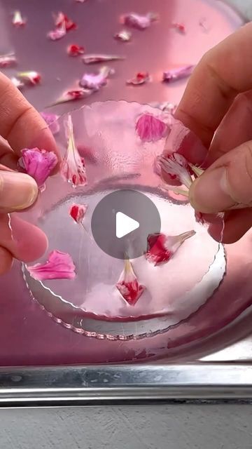 two hands are touching petals in a glass bowl with pink flowers on the bottom and one hand is reaching for it