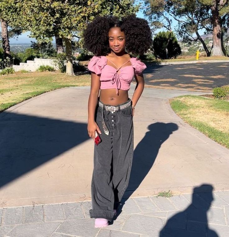 a woman with an afro standing in front of a tree and walkway wearing grey pants