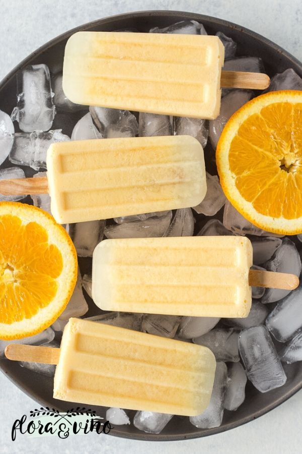 four popsicles with orange slices and ice cubes on a black plate surrounded by ice