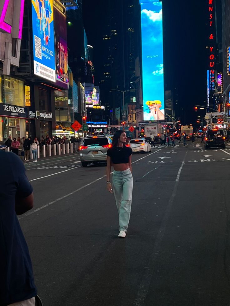 a woman walking down the middle of a busy city street at night with tall buildings in the background