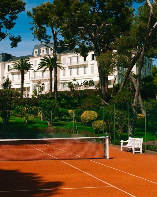 a tennis court in front of a large white building with trees on the other side