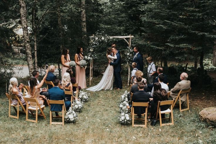 a wedding ceremony in the woods with people sitting on chairs and looking at each other