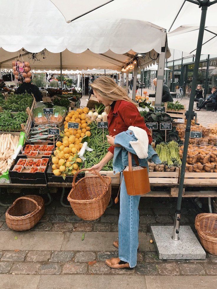 a woman standing in front of a fruit and vegetable stand holding a brown bag with her right hand