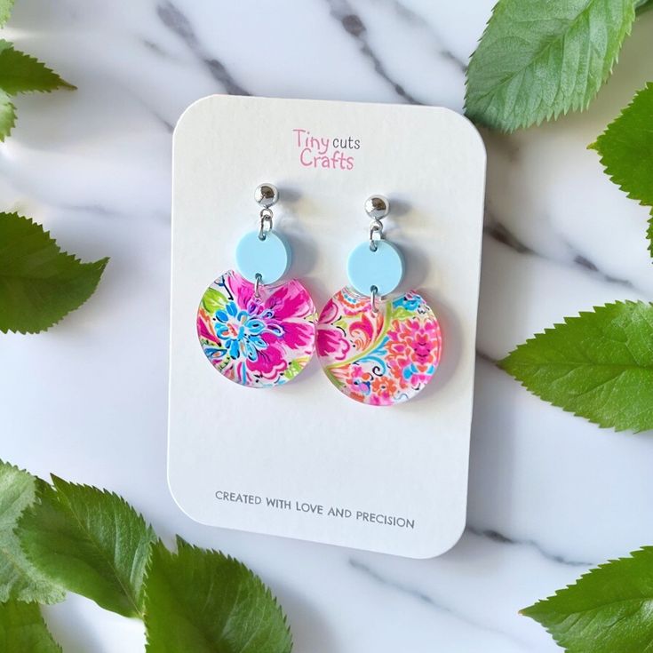 a pair of colorful earrings on top of a white table next to green leafy leaves