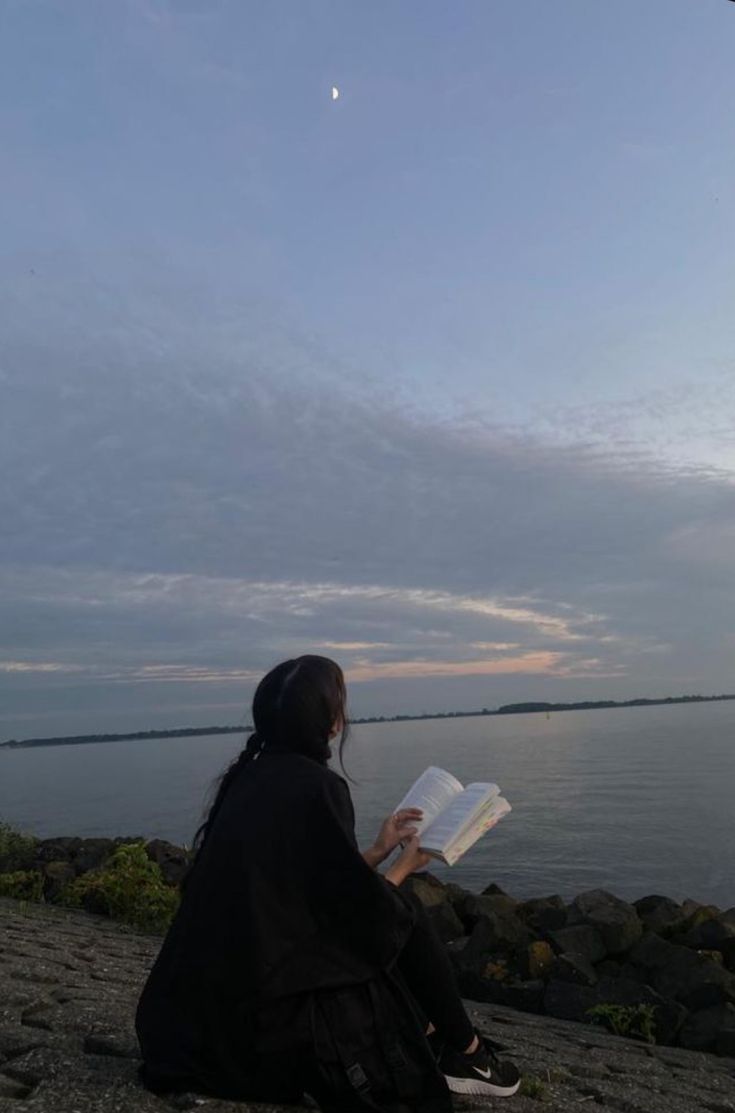 a woman sitting on the beach reading a book while looking at the sky and water