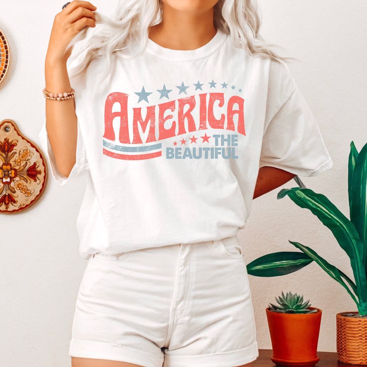 Celebrate the 4th of July in our retro America The Beautiful graphic t-shirt. Perfect for the beach, summer BBQs and all your 4th of July activities.  Comfort Colors introduces its garment-dyed t-shirt 100% with ring-spun cotton. The soft-washed, garment-dyed fabric brings extra coziness to your wardrobe while the relaxed fit makes it an excellent daily choice. The double-needle stitching throughout the tee makes it highly durable while the lack of side-seams helps the shirt retain its tubular s Retro American Flag Print Summer T-shirt, American Flag Print Cotton Top For Beach, American Retro Graphic Print Summer Tops, White American Flag Print Top For The Beach, White American Flag Print Top For Beach, White American Flag Print Tops For Beach, American Flag Print Summer Beach Top, White Tops With American Flag Print For Beach, White Retro Top For 4th Of July