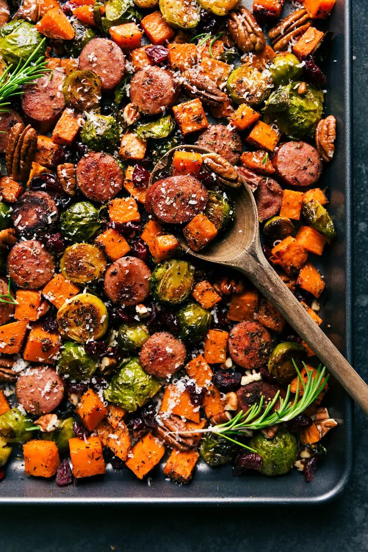 a tray filled with sausage, sweet potatoes and brussel sprouts next to a wooden spoon