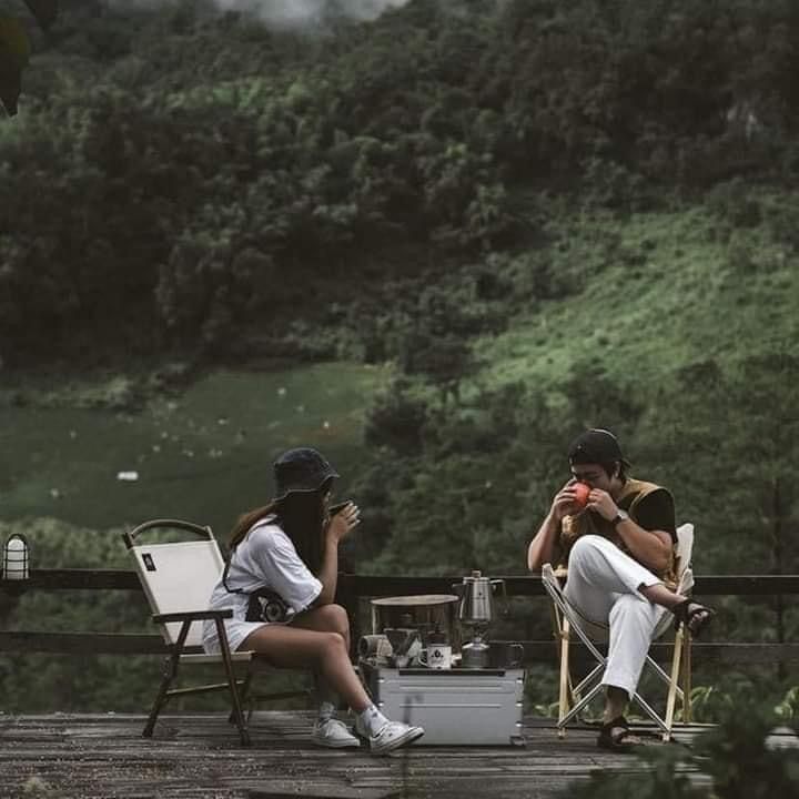 two people sitting on chairs in the middle of a forest, one is taking a photo