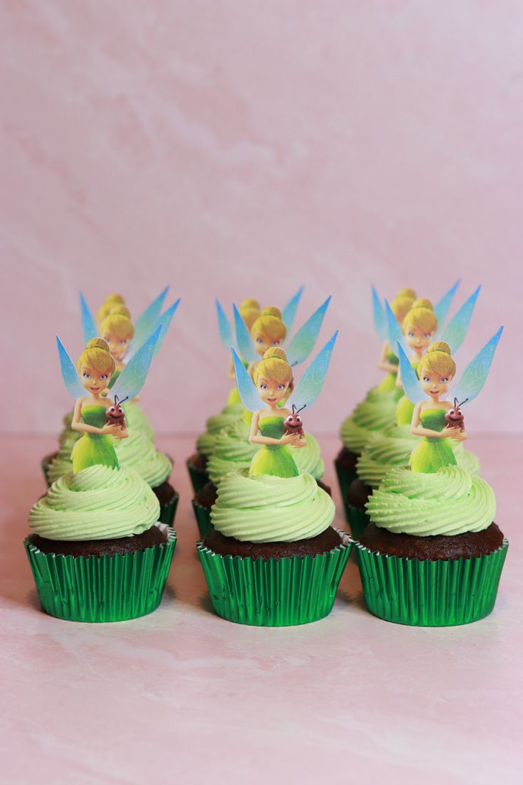 four cupcakes with green frosting and tinker fairy on top, sitting in front of a pink background