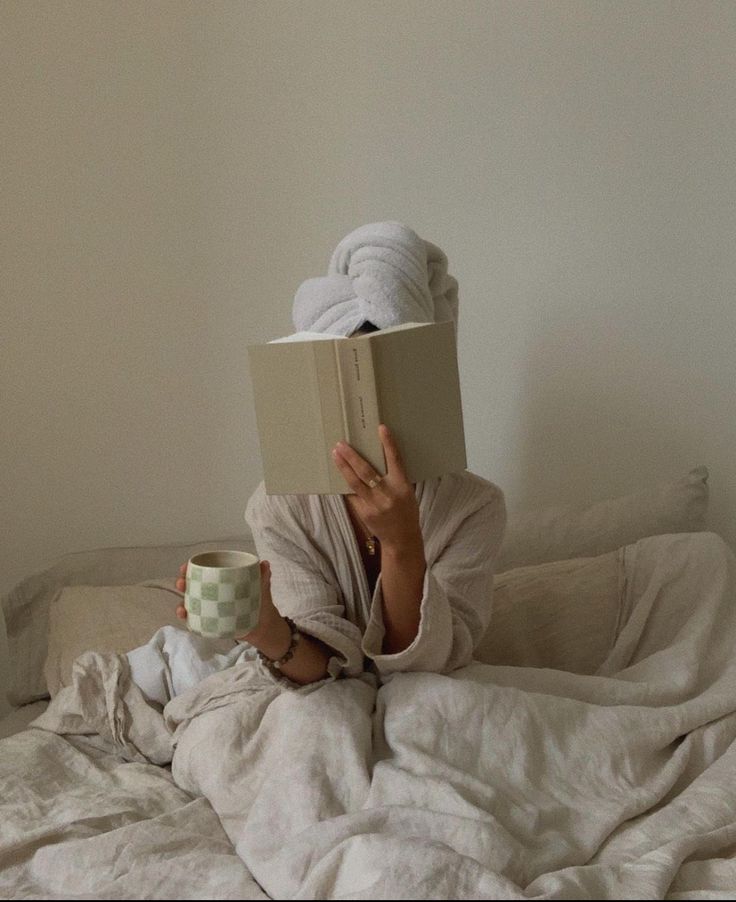 a person laying in bed reading a book and holding a cup