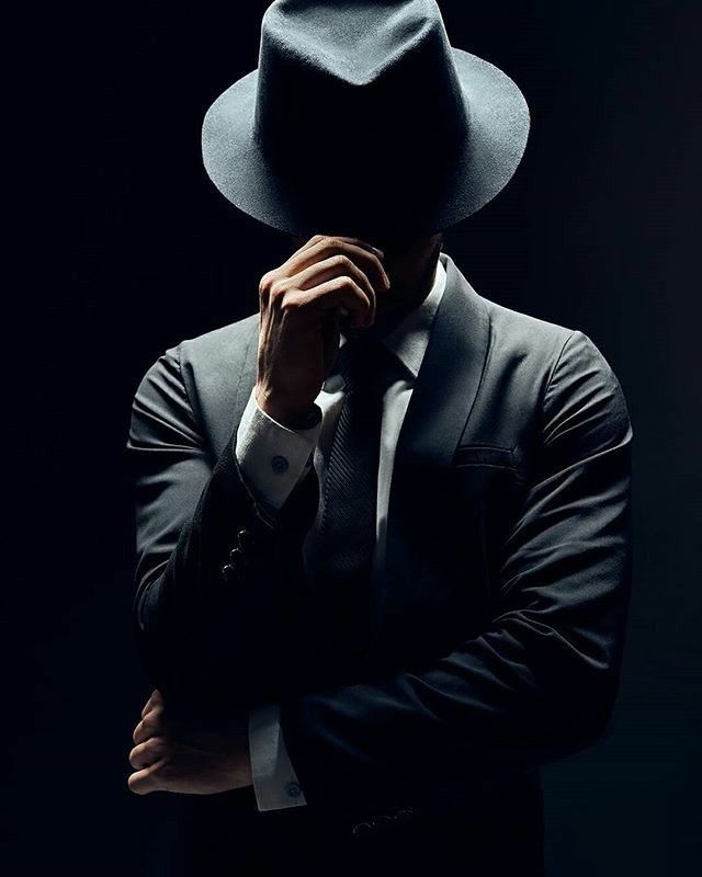 a man in a black suit and hat is holding his hands to his face while standing against a dark background