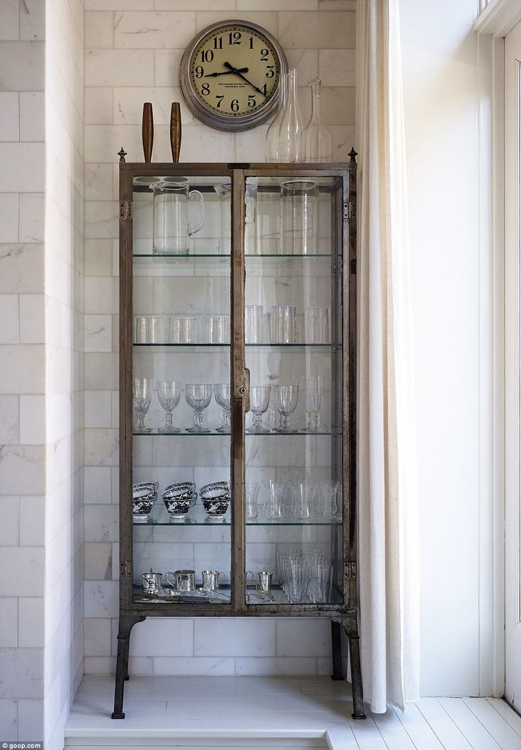 an old china cabinet with wine glasses and a clock on the wall next to it