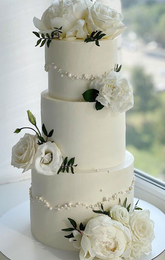 a wedding cake with white flowers and pearls on the bottom tier is sitting in front of a window