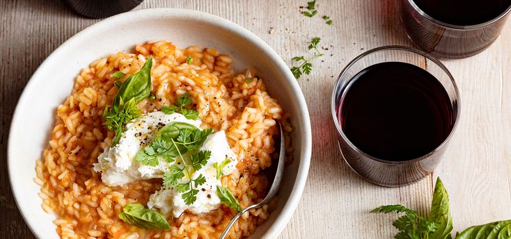 a white bowl filled with pasta next to two glasses of wine and herbs on a table