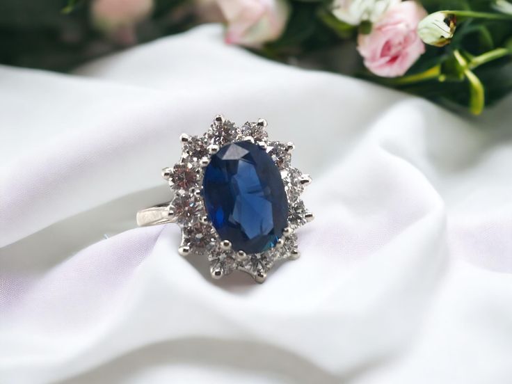 an oval shaped blue stone surrounded by small white diamonds on a white satin with pink flowers in the background