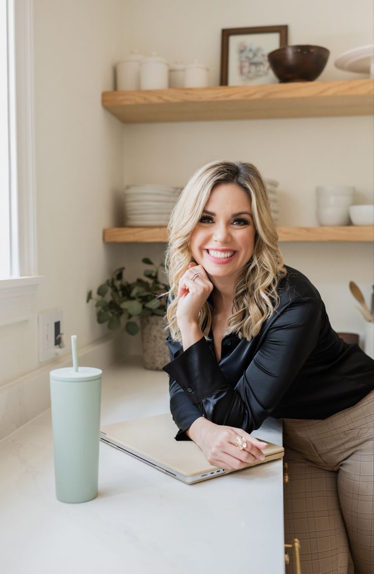 a woman sitting at a kitchen counter with a laptop in front of her, smiling