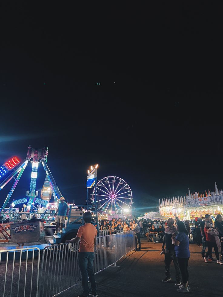 people are standing near a fence at night with ferris wheel in the background and carnival rides on the other side