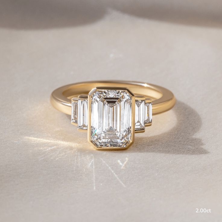 an emerald cut diamond ring with three baguets on the band, set in yellow gold