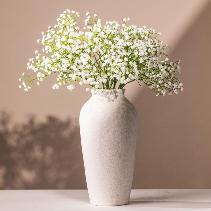 a white vase with flowers in it sitting on a table next to a pink wall